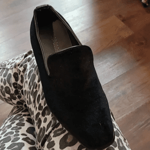 A black suede loafer shoe on a person's lap with leopard print pants.